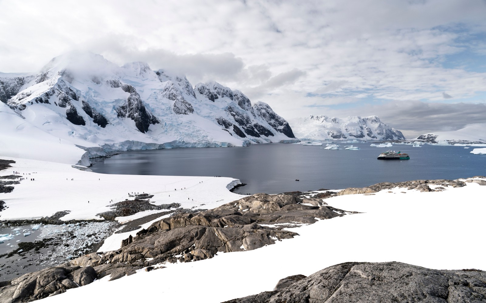 An expedition ship at anchor in a snowy Antarctic bay below glaciated mountains.