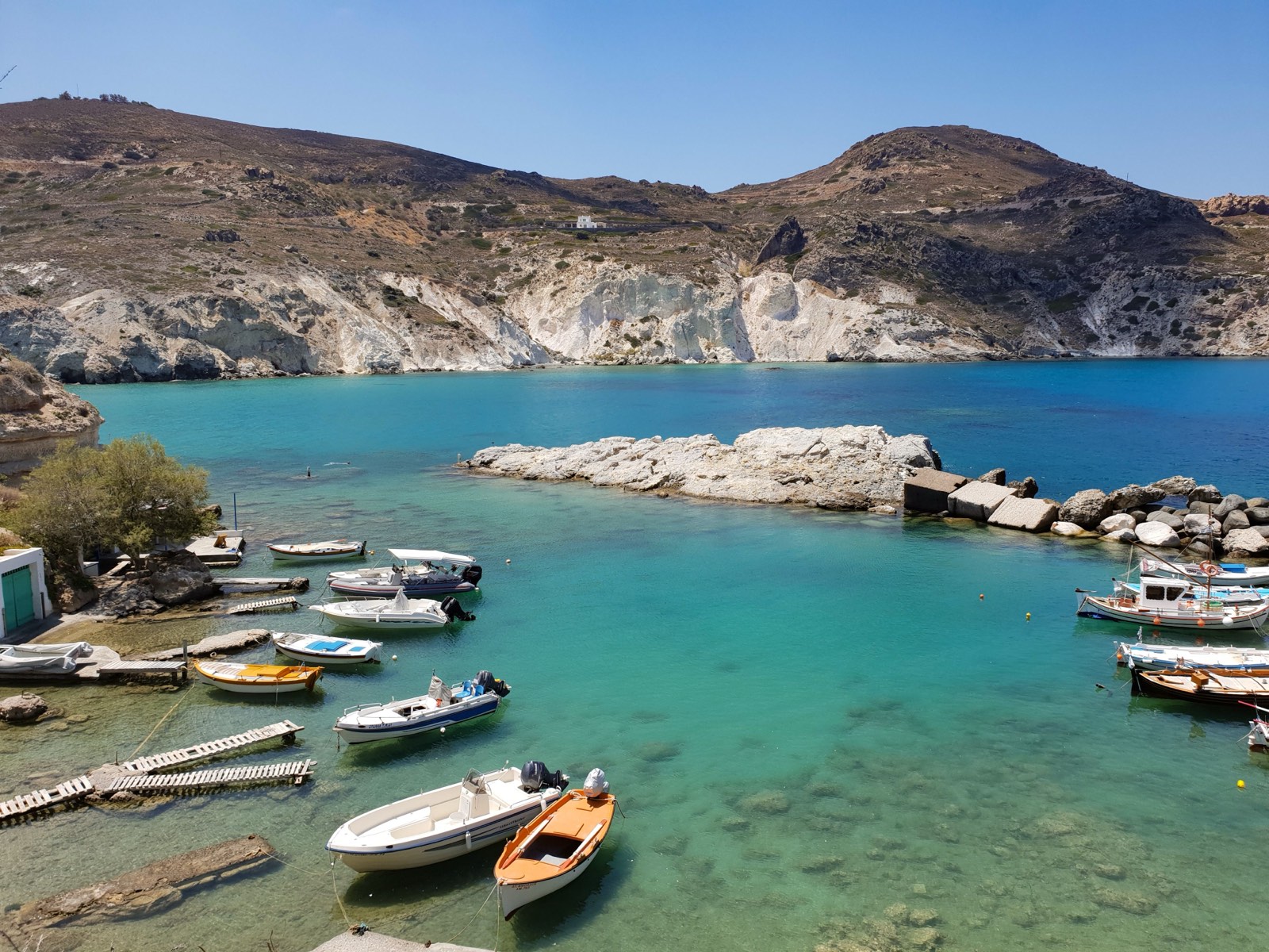 A turquoise cove on Milos with small fishing boats moored below pale volcanic cliffs.