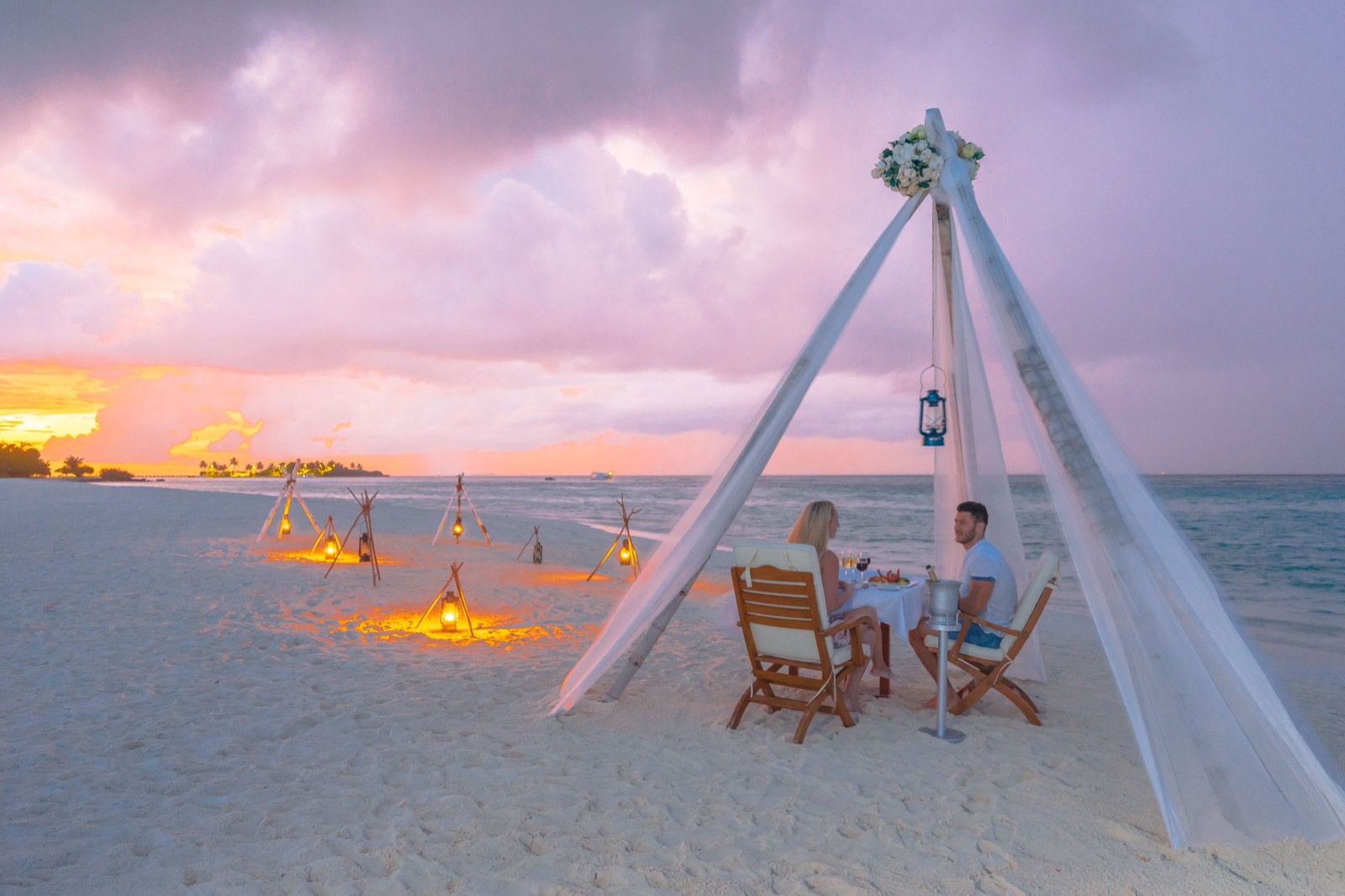 A dinner table for two under a white draped canopy on a beach at sunset, lanterns set in the sand.