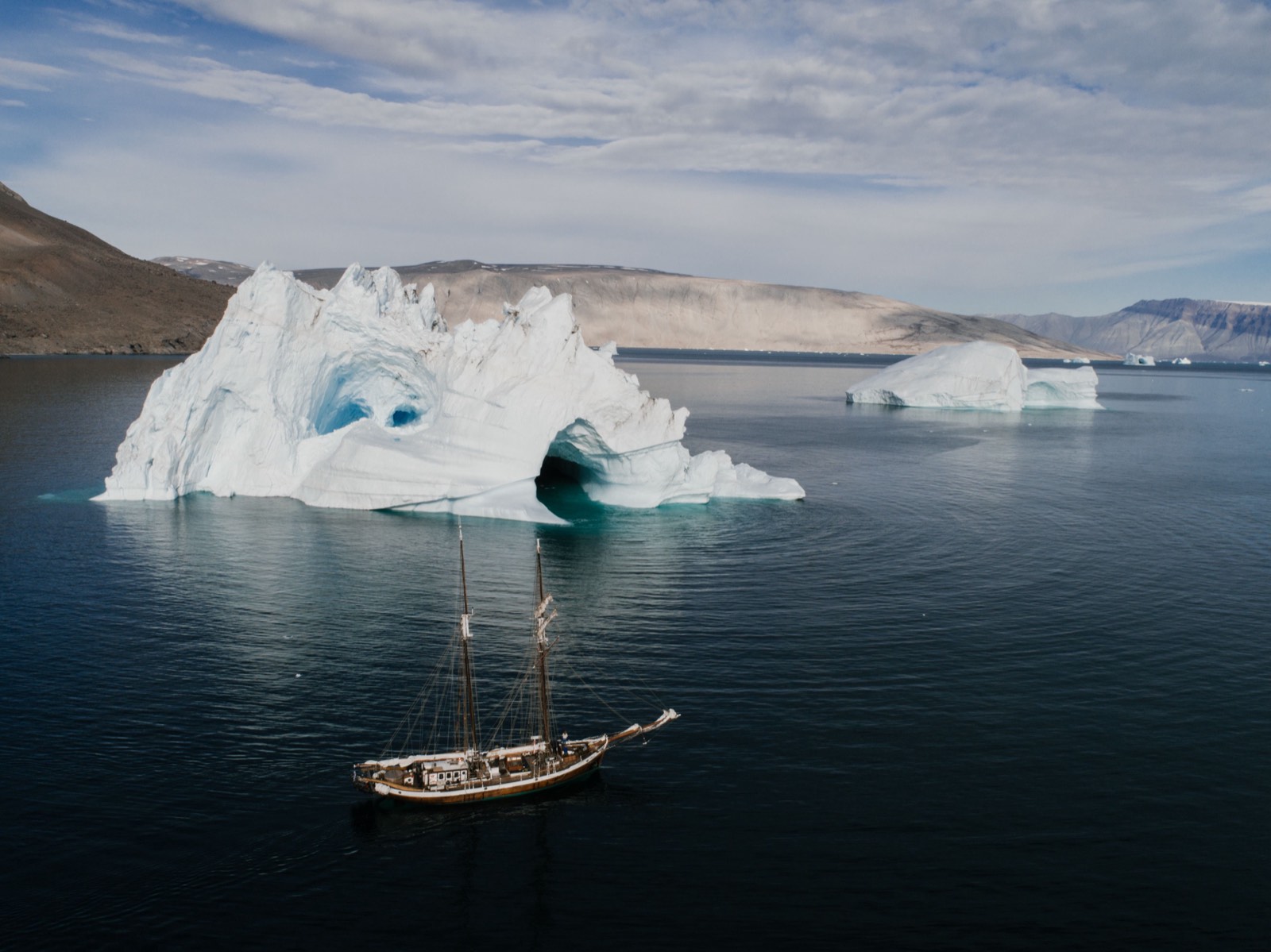 A two-masted sailing ship beside a large sculpted iceberg in calm, dark water, with bare hills behind.