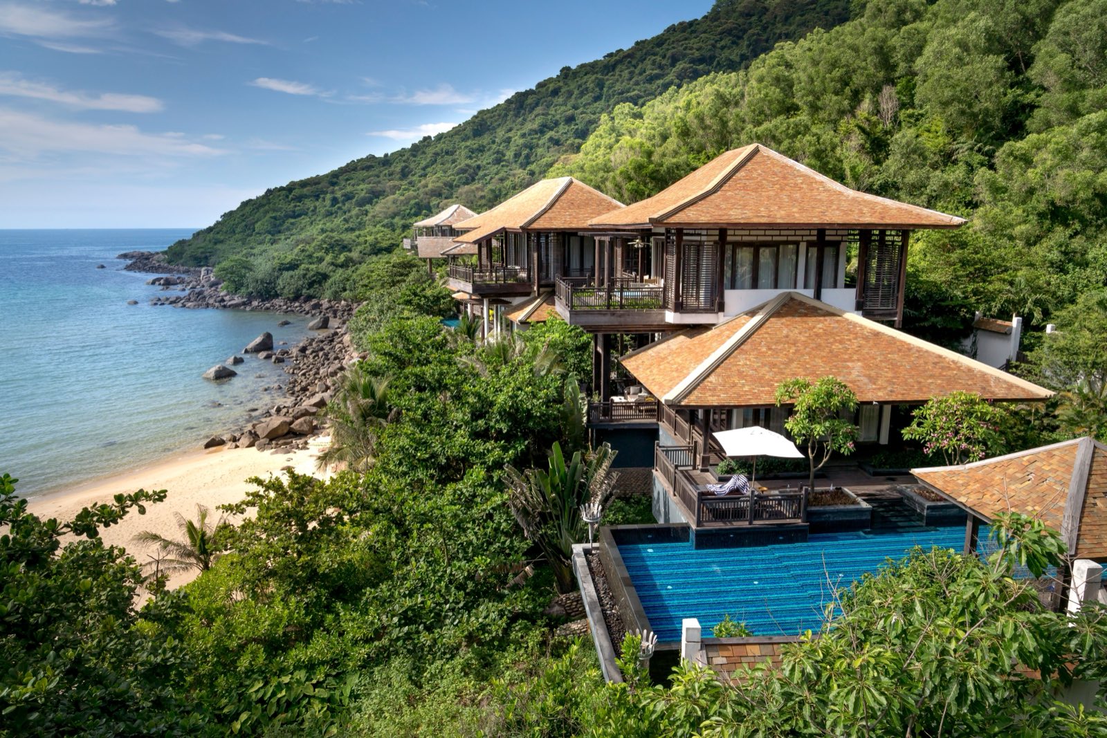 A tiered villa with tiled roofs and a private pool on a forested hillside above a small sandy beach.